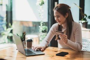 jovem mulher asiática sorridente descansando e fazendo compras on-line em casa, mulher feliz usando laptop e cartão de crédito para compras on-line. conceito de pagamento online. foto