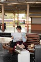 feliz jovem viajante asiática ou mochileiro usando mapa escolher para onde viajar com bagagem na estação de trem, conceito de viagem de férias de verão foto