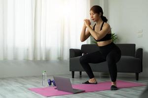 ajuste o exercício de treino online desportivo jovem asiático em casa. menina saudável ativa desfrutar de esporte pilates yoga treinamento de fitness no computador portátil alongamento no tapete de ioga assistindo aula de vídeo foto