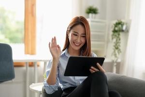 imagem de mulher asiática feliz sorrindo e acenando com a mão no tablet digital, enquanto fala ou conversa em videochamada em casa foto