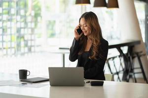 jovem mulher asiática caridosa usando seu laptop enquanto ligava para o telefone e sentada em seu local de trabalho. foto