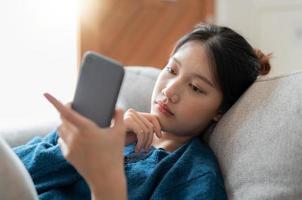 alegre jovem mulher asiática usando telefone celular enquanto está sentado em um sofá em casa. foto