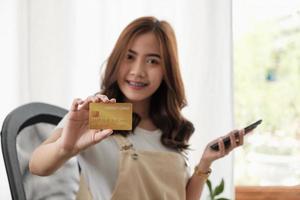 jovem e bela mulher segurando o cartão de crédito enquanto estiver usando smartphone, compras de pagamento on-line foto