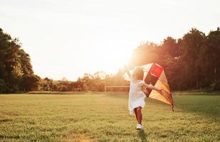 portões de futebol atrás. garota feliz em roupas brancas divirta-se com pipa no campo. natureza bela foto