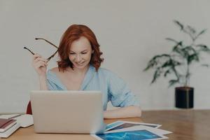 mulher ocupada positiva com aparência europeia trabalha em projeto digital freelance, ganha dinheiro online em trabalho à distância, senta-se no local de trabalho com papéis e bloco de notas, veste camisa casual, segura óculos foto