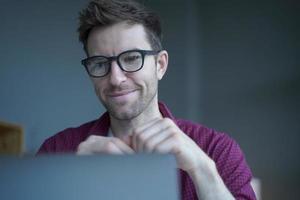 sorridente jovem alemão freelancer em óculos trabalhando remotamente on-line no laptop do escritório em casa foto