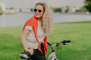 conceito de estilo de vida ativo. jovem positiva com cabelos claros encaracolados, anda de bicicleta no gramado verde, usa óculos escuros, camiseta branca e suéter vermelho nos ombros, carrega bolsinha, estando de bom humor foto