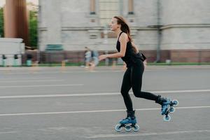 conceito de patins e fitness de verão. caber a jovem mulher de cabelos escuros que gosta de andar de patins exercita todas as poses do corpo na estrada ao longo do asfalto durante o dia ensolarado usa roupas confortáveis tem figura esportiva foto