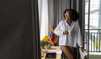 garota afro-americana com bandeira do arco-íris lgbtq em seu quarto de cama para sair do armário e celebração do mês do orgulho para promover a diversidade sexual e igualdade no conceito de orientação homossexual foto