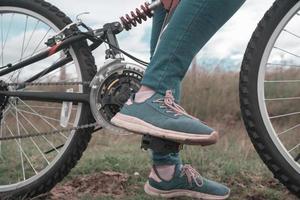 detalhe dos pés de uma jovem vestindo calça azul nos pedais de sua bicicleta no meio do campo em um dia nublado foto