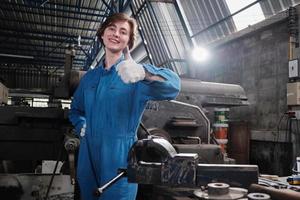 um trabalhador da indústria feminina caucasiano jovem alegre em uniforme de segurança relaxa e polegares para cima após o sucesso do trabalho e expressa a felicidade do sorriso em uma fábrica mecânica. profissão de engenheiro. foto