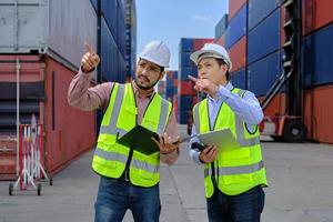 dois trabalhadores asiáticos profissionais em uniformes de segurança e capacetes trabalham em um terminal logístico com muitas pilhas de contêineres, carregando mercadorias de transporte de controle para a indústria de transporte de carga. foto