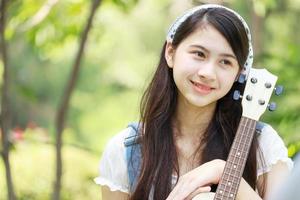menina asiática tocando ukulele e sorrindo alegremente no parque. foto