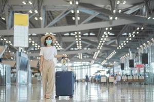 jovem mulher usando máscara facial com bagagem andando no aeroporto, proteção contra infecção por doença de coronavírus, viajante asiática com chapéu. hora de viajar após o conceito de dose de reforço de vacina