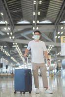 jovem homem usando máscara facial com bagagem andando no aeroporto, proteção contra infecção por doença de coronavírus, viajante asiático com chapéu. hora de viajar após o conceito de dose de reforço de vacina foto