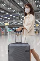 jovem mulher usando máscara facial com bagagem verificando o tempo de voo no aeroporto, proteção contra infecção por doença de coronavírus, viajante asiática pronta para viajar. novo normal e viajar sob covid-19