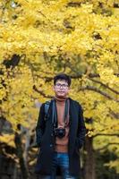 homem feliz desfruta no parque ao ar livre na temporada de outono, viajante asiático de casaco e câmera contra fundo de folhas de ginkgo amarelo foto