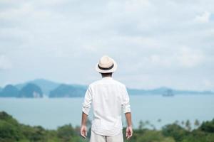 homem viajante feliz na camisa branca desfrutar de uma bela vista, turista sozinho com chapéu em pé e relaxando sobre o oceano. conceito de viagens, verão e férias foto