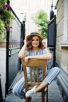 retrato de uma jovem fabulosa em macacão listrado e chapéu sentado e posando na cadeira ao ar livre.