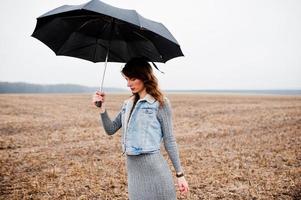 retrato de menina morena encaracolada na jaqueta jeans com guarda-chuva preto no campo. foto