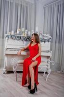 mulher linda elegante no vestido de noite vermelho longo sentado no estúdio contra piano com velas decoração de ano novo. foto