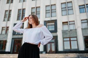 retrato de uma jovem fabulosa bem sucedida na blusa branca e calças pretas largas posando nas escadas com um enorme edifício branco no fundo. foto