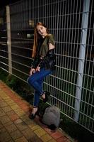 retrato noturno do desgaste do modelo de menina em jeans e jaqueta de couro contra cerca de ferro. foto
