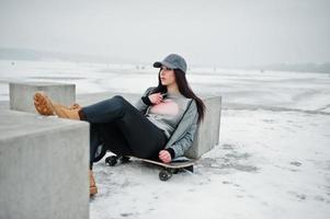 menina morena elegante boné cinza, estilo casual de rua com skate em dia de inverno. foto