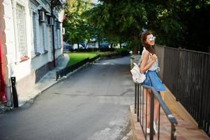 garota elegante encaracolada usa saia jeans azul, blusa com bolsa. foto