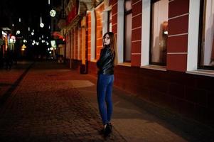 retrato noturno do modelo de menina usar óculos, jeans e jaqueta de couro, com mochila, contra as ruas da cidade. foto
