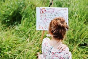 retrato de uma linda jovem feliz em lindo vestido sentado na grama e pintando em papel com aquarelas. foto