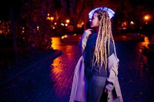 menina com dreadlocks andando na rua à noite da cidade contra luzes de guirlanda. foto