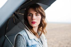 retrato de menina morena encaracolada na jaqueta jeans com guarda-chuva preto no campo. foto