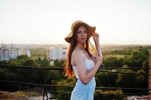 retrato de uma bela jovem de camiseta branca e saia azul posando no telhado com seu chapéu laranja ao pôr do sol. foto