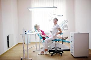 linda dentista feminina posando e sorrindo com seu adorável paciente deitado em uma cadeira odontológica com vestido vermelho-violeta. foto