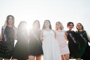 grupo de 7 meninas usam preto e 2 noivas na festa de despedida contra a praia ensolarada. foto