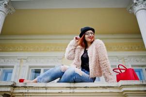 garota ruiva de óculos e chapéu com bolsa vermelha posou perto de casa vintage. foto