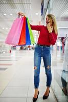 retrato de uma jovem deslumbrante de blusa vermelha, jeans casuais rasgados e salto alto posando com sacolas de compras no shopping. foto