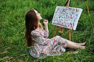 retrato de uma linda jovem feliz em lindo vestido sentado na grama e pintando em papel com aquarelas. foto