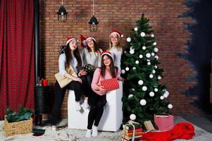 quatro amigas fofas usam blusas quentes, calças pretas e chapéus de papai noel contra a árvore de ano novo com decoração de natal no estúdio. foto