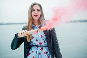 garota elegante na jaqueta de couro segure o clarão de fumaça rosa no dia de inverno contra o lago congelado. foto