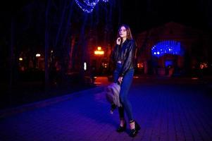 retrato noturno do modelo de menina usar óculos, jeans e jaqueta de couro, com mochila nas mãos, contra guirlanda de luzes azuis da rua da cidade. foto