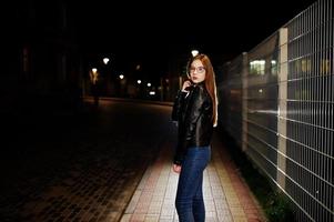 retrato noturno do desgaste do modelo de menina em jeans e jaqueta de couro contra cerca de ferro. foto