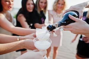 grupo de 8 meninas usam preto e 2 noivas na festa de despedida contra praia ensolarada bebendo champanhe. foto