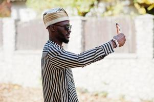 retrato de homem afro-americano preto elegante no chapéu e óculos de sol contra fundo de outono ensolarado outono fazendo selfie do telefone celular. pessoas ricas da áfrica em trajes tradicionais. foto