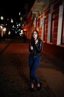 retrato noturno do modelo de menina usar óculos, jeans e jaqueta de couro, com mochila, contra as ruas da cidade. foto