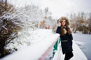 linda menina morena em roupas quentes de inverno. modelo na jaqueta de inverno contra o lago congelado no parque. foto