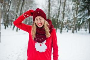 loira de cachecol vermelho, chapéu e suéter de papai noel posando no parque em dia de inverno. foto