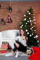 linda garota usa blusas quentes, calças pretas contra árvore de ano novo com decoração de natal no estúdio. foto