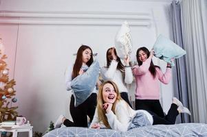 quatro amigas fofas usam blusas quentes e calças pretas na cama no quarto decorado de ano novo no estúdio, brincam com travesseiros. foto